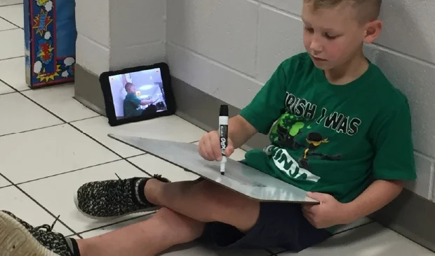 Boy sitting on floor writing on eraser board while recording video on tablet
