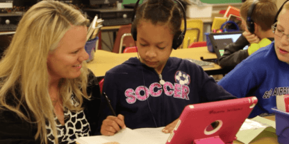 A teacher kneeling down to provide support to students working on iPads.