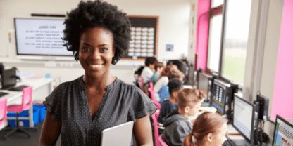 A teacher standing in the foreground while students work on computers in the background.