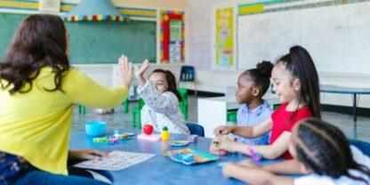 A teacher giving a high-five to her student across a small desk while other students look on.