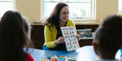 A teacher holding a colorful shape identification sheet up for students on the other side of the table.