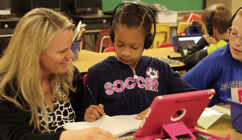 A teacher kneeling over a desk to help two students with iPads