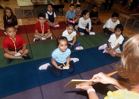 Students sitting on a mat watching their teacher read a book